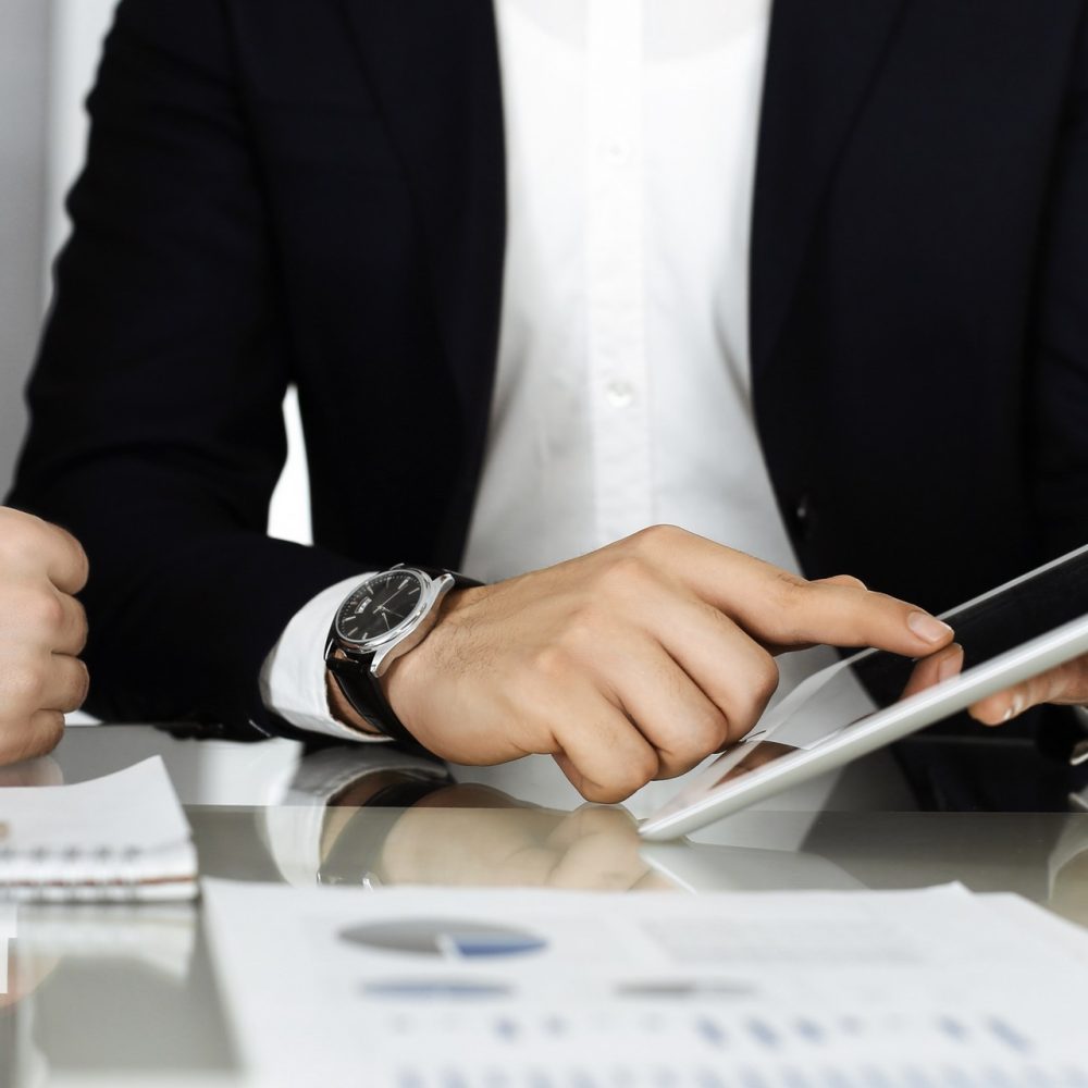 Business people using tablet computer while working together in modern office. Unknown businessman sitting at the desk with his colleague or partner. Teamwork and partnership concept.